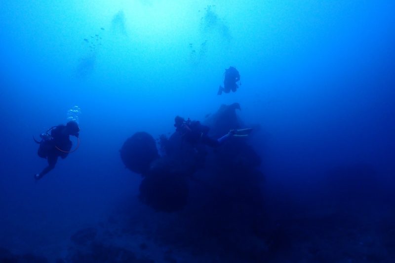 青い海の中に沈んでいる巨大な沈没船のスクリューを3人のダイバーが観察している。