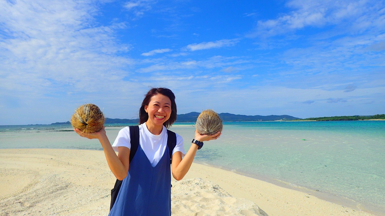 両手にヤシの実を持った笑顔の女性が、海を背景に写っている