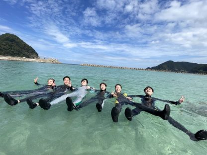 ビーチでWETスーツを着たダイバー6人が手をつないで浮かんでいる様子